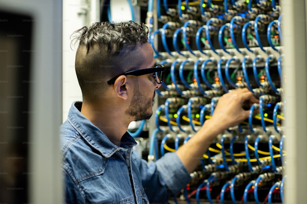 Ernsthafter junger arabischer Mainframe-Ingenieur mit Brille, der am Serverschrank steht und das Datenbankspeichersystem analysiert