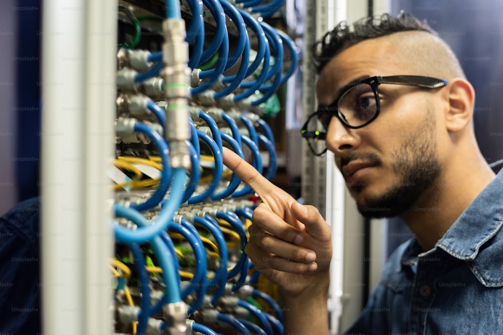 Imagem cortada de um jovem engenheiro de rede do Oriente Médio pensativo sério com barba apontando para o cabo e verificando-o na sala do servidor