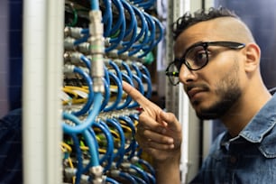 Imagen recortada de un joven ingeniero de redes de Oriente Medio serio y pensativo con barba apuntando al cable y revisándolo en la sala de servidores