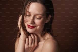 Concepto de belleza y maquillaje. Retrato de cerca de una joven pecosa sensual y sonriente morena que cierra los ojos tocando su cabello. Aislado en marrón con espacio de copia a la derecha