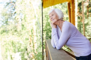 Happy serne senior female in casualwear enjoying rest in the country house while standing by wooden banisters