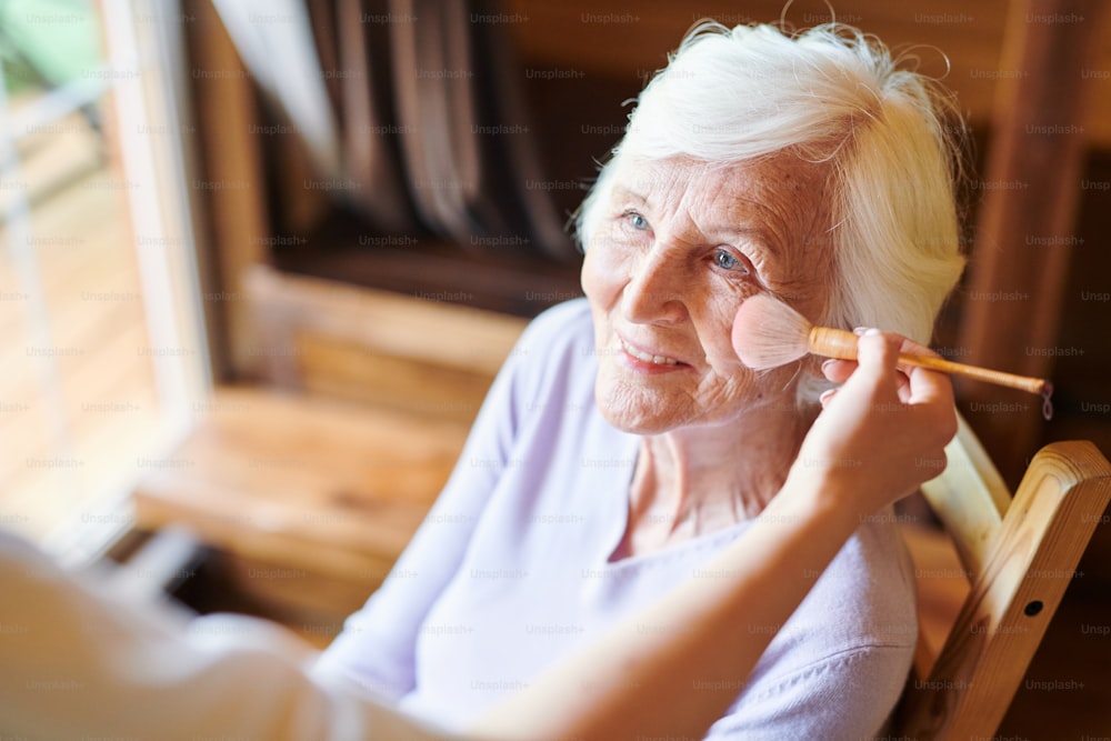 Happy senior female with short white hair looking at beautician during makeup procedure in beauty salon