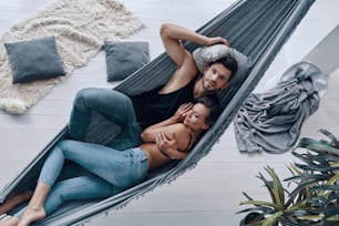 Top view of beautiful young couple embracing and smiling while resting in the hammock indoors