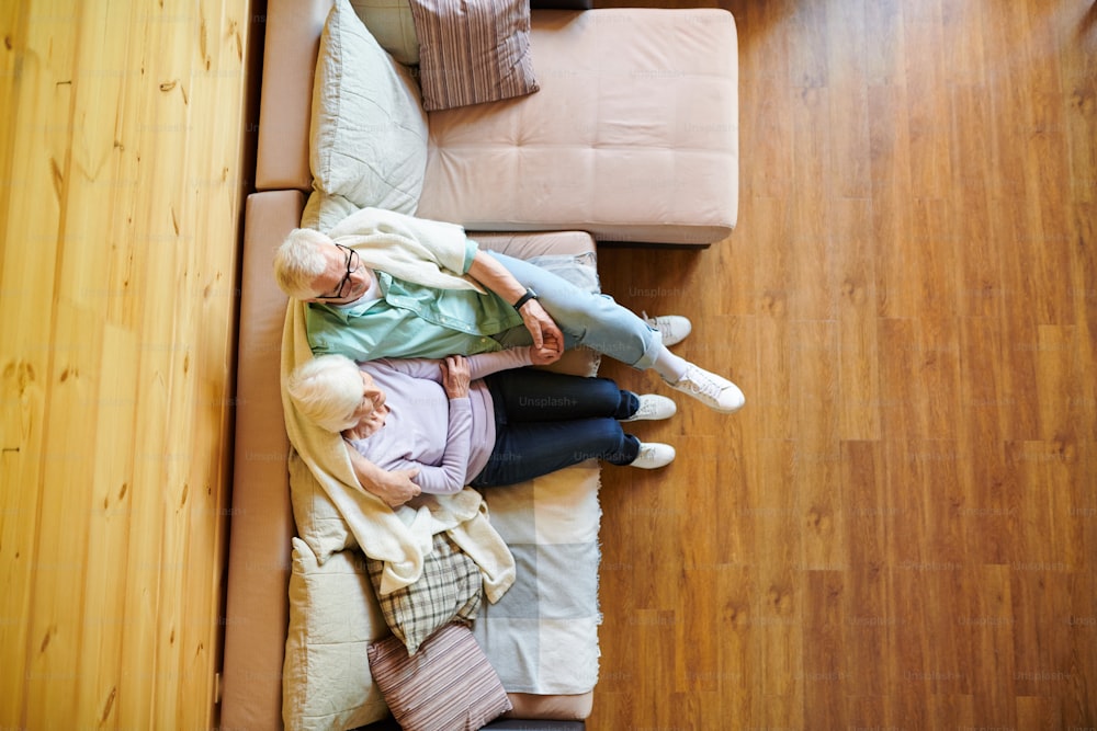 Overview of restful senior man and woman in casualwear sitting on couch in front of tv set while relaxing in their country house