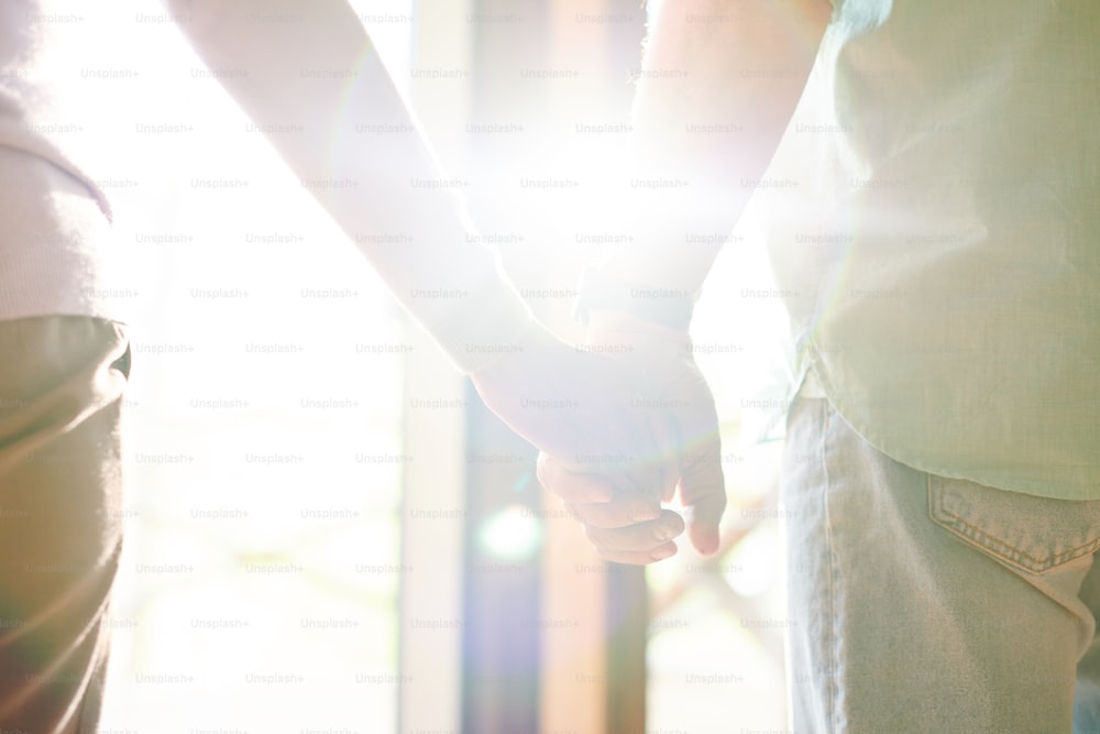 Flare of sunlight between hands of casual senior spouses standing in front of window on sunny day