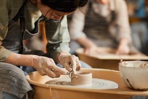 陶器の車輪に座り、濡れた粘土容器を形作りながら工芸道具を使うエプロンを着た集中したアジアの職人の接写