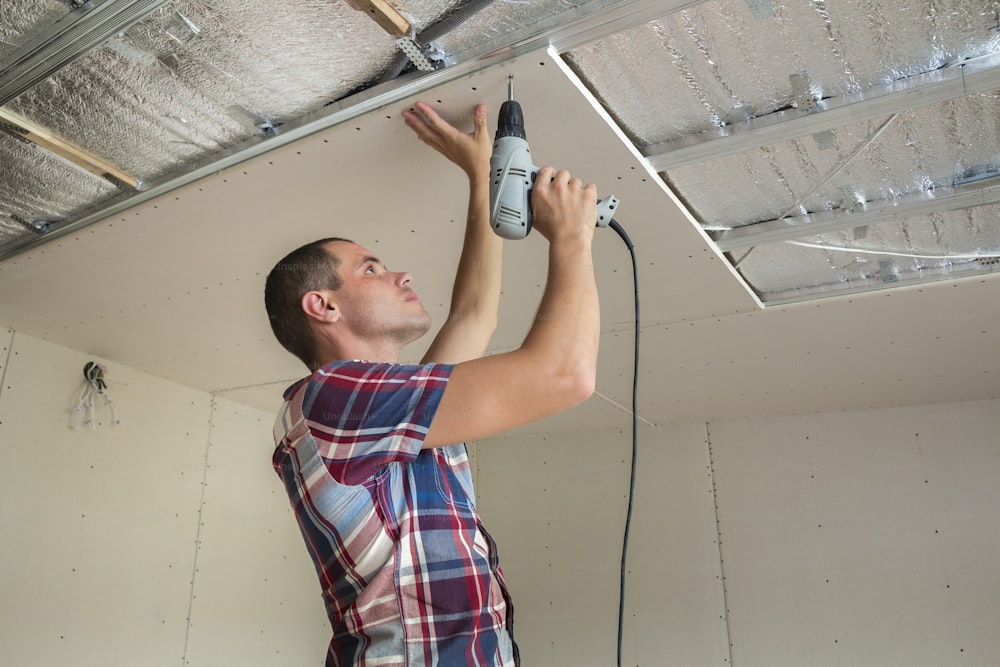 Young man in usual clothing and work gloves fixing drywall suspended ceiling to metal frame using electrical screwdriver on ceiling insulated with shiny aluminum foil. DIY, do it yourself concept.