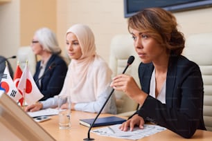 Young confident female speaker in formalwear keeping mike by her mouth while going to answer or ask question at conference