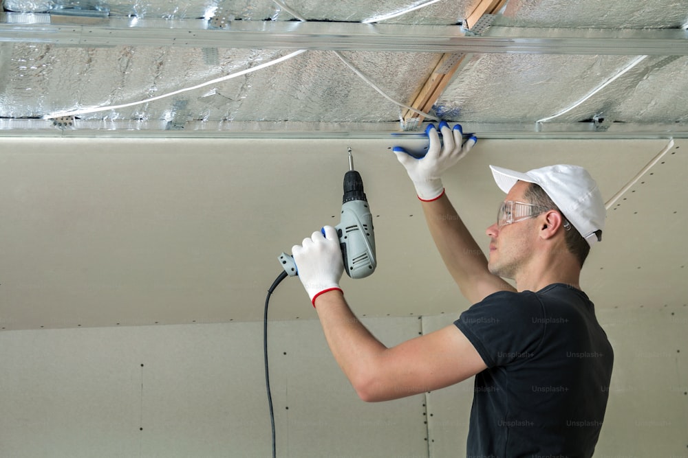 光沢のあるアルミホイルで断熱された天井の電気ドライバーを使用して、乾式壁の吊り天井を金属フレームに固定するゴーグルの若い男性。リノベーション、建設、自分でコンセプトをやる。