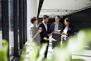 Group of content enterprising business colleagues in formalwear standing in lobby and viewing papers while discussing ideas for new project