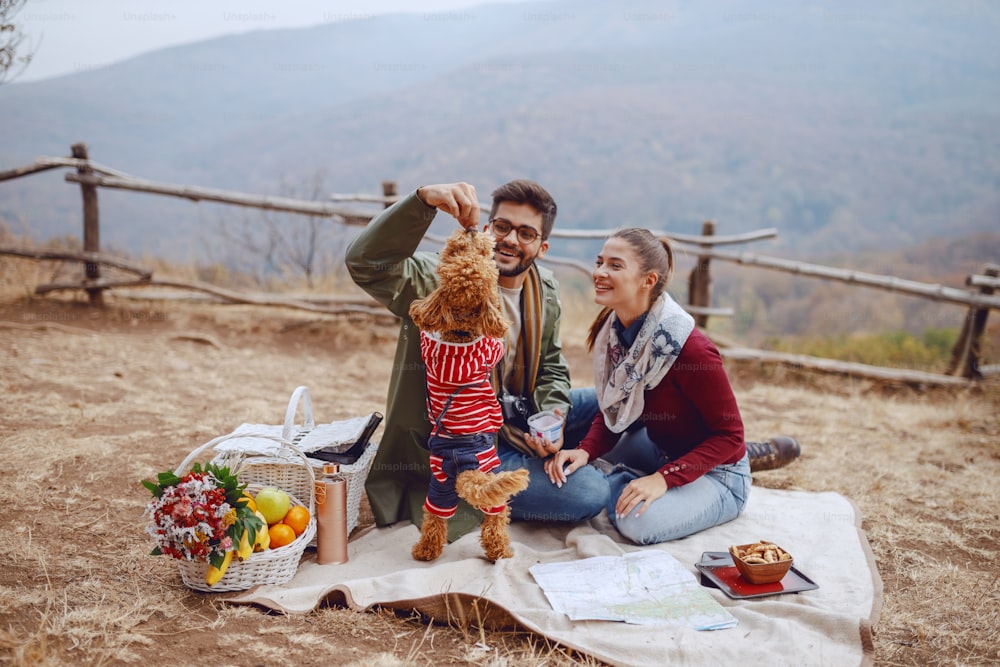 Attractive multicultural couple sitting on blanket and playing with their dog. Picnic at autumn concept.