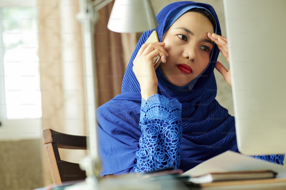 Mulher muçulmana estressada frustrada falando ao telefone ao ler e-mail na tela de seu computador