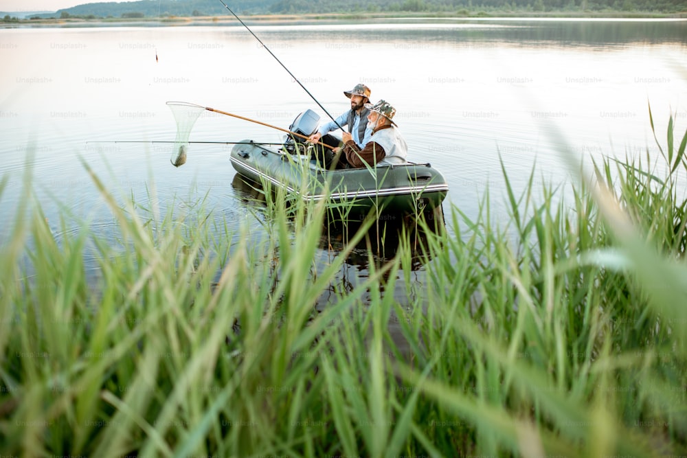 Grandfather with adult son fishing on the inflatable boat on the lake with green cane on the foreground early in the morning