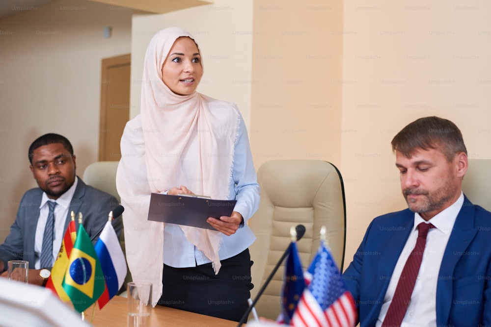 Joven musulmana con hiyab haciendo un informe en la conferencia mientras está de pie frente a la audiencia en la sala