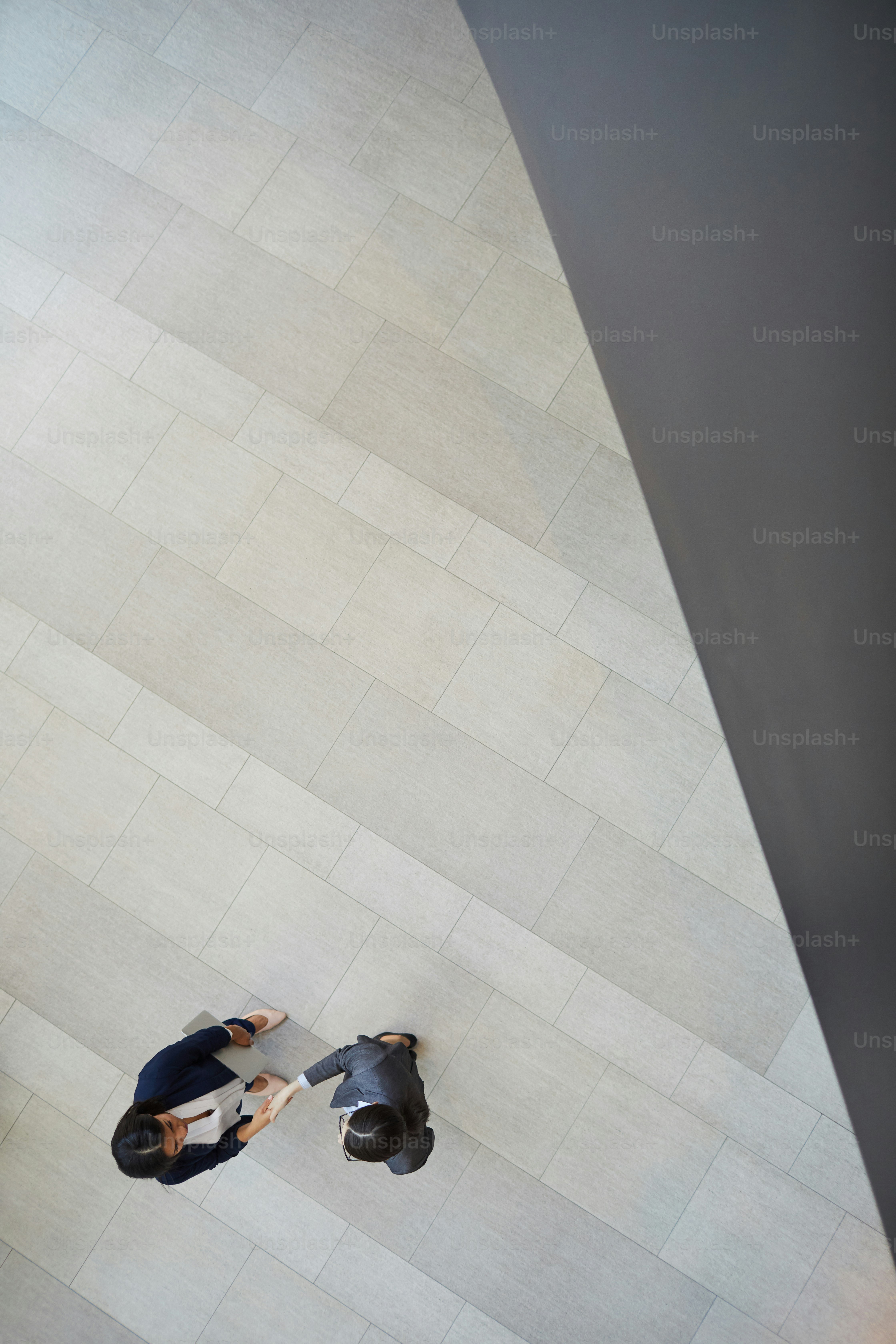 Vue en grand angle de femmes d’affaires multiethniques debout dans le hall du bureau avec un sol carrelé gris et se serrant la main après la conclusion de l’accord