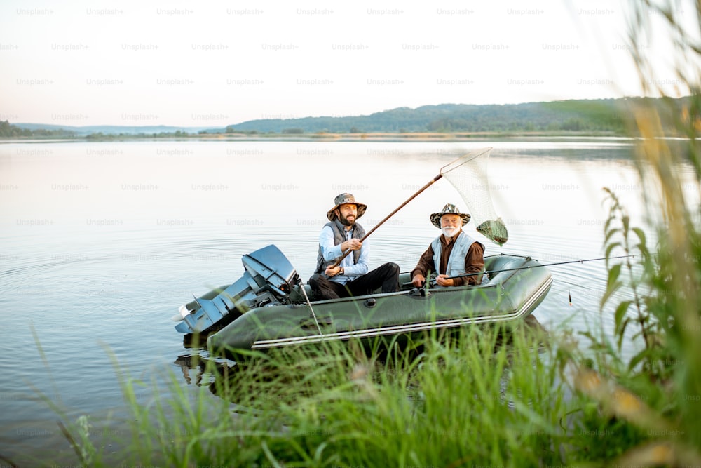 Abuelo con hijo adulto pescando en el bote inflable en el lago con caña verde en primer plano temprano en la mañana