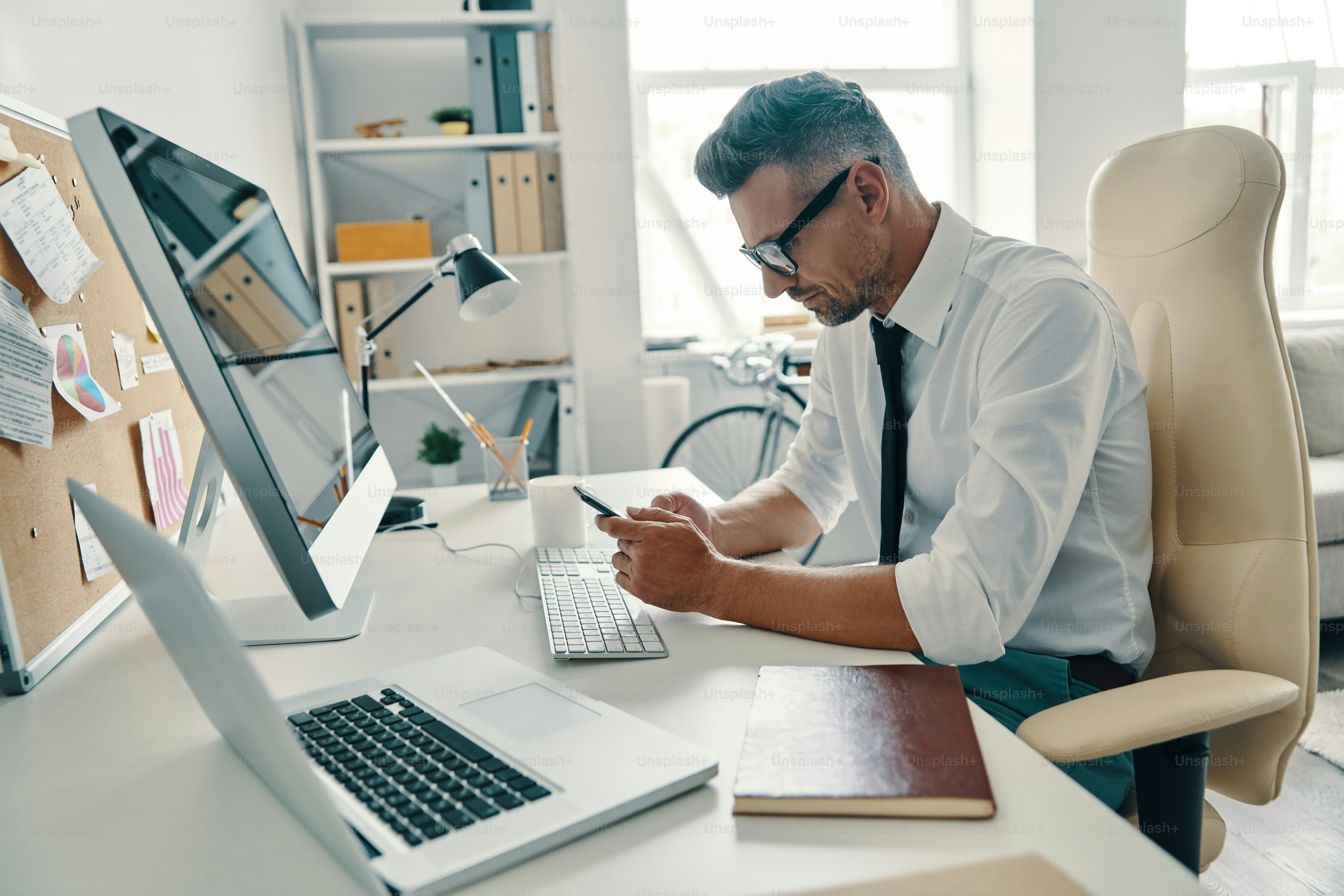 Thoughtful young man in shirt and tie using smart phone while sitting in the office
