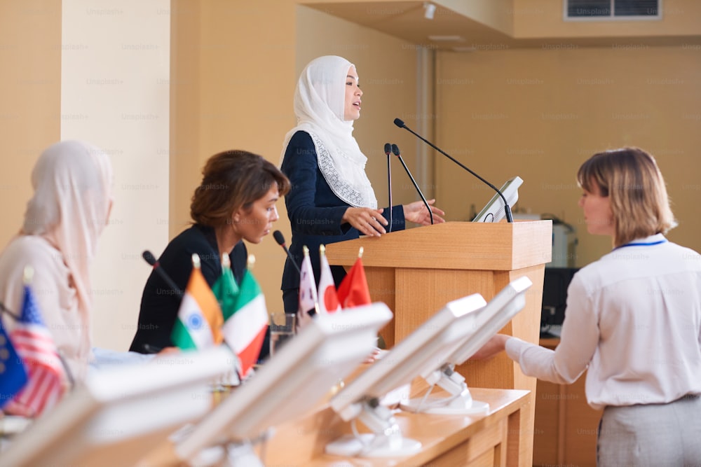 Giovane delegata contemporanea in hijab e giacca e cravatta che parla al microfono mentre è in piedi vicino alla tribuna alla conferenza politica