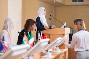 Young contemporary female delegate in hijab and suit speaking in microphone while standing by tribune at political conference