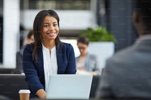 Happy confident young black woman in jacket sitting in front of laptop and talking to business partner while holding meeting in cafe