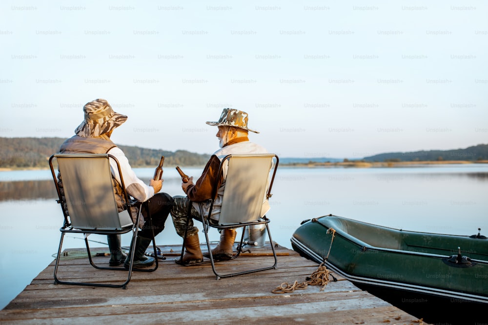 Avô com filho adulto apreciando cerveja, sentados juntos no cais enquanto pescam no lago no início da manhã