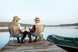 Grandfather with adult son enjoying beer, sitting together on the pier while fishing on the lake early in the morning