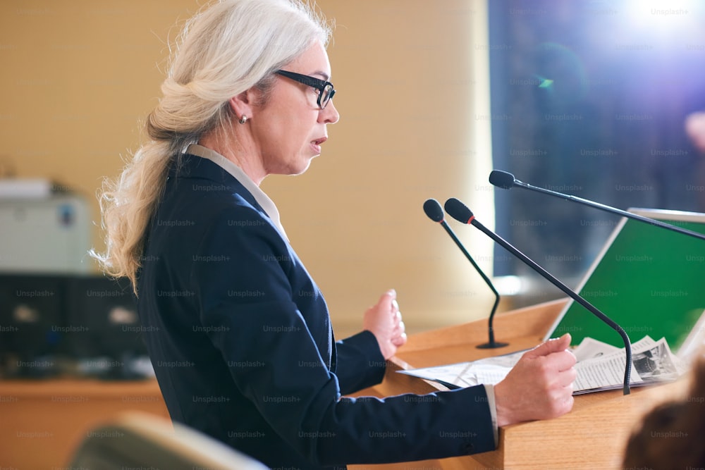 Delegada madura e elegante de terno falando ao microfone enquanto está ao lado da tribuna na sala de conferências na cúpula