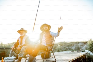 Grandfather with adult son fishing together on the wooden pier during the morning light. View from the side of the lake