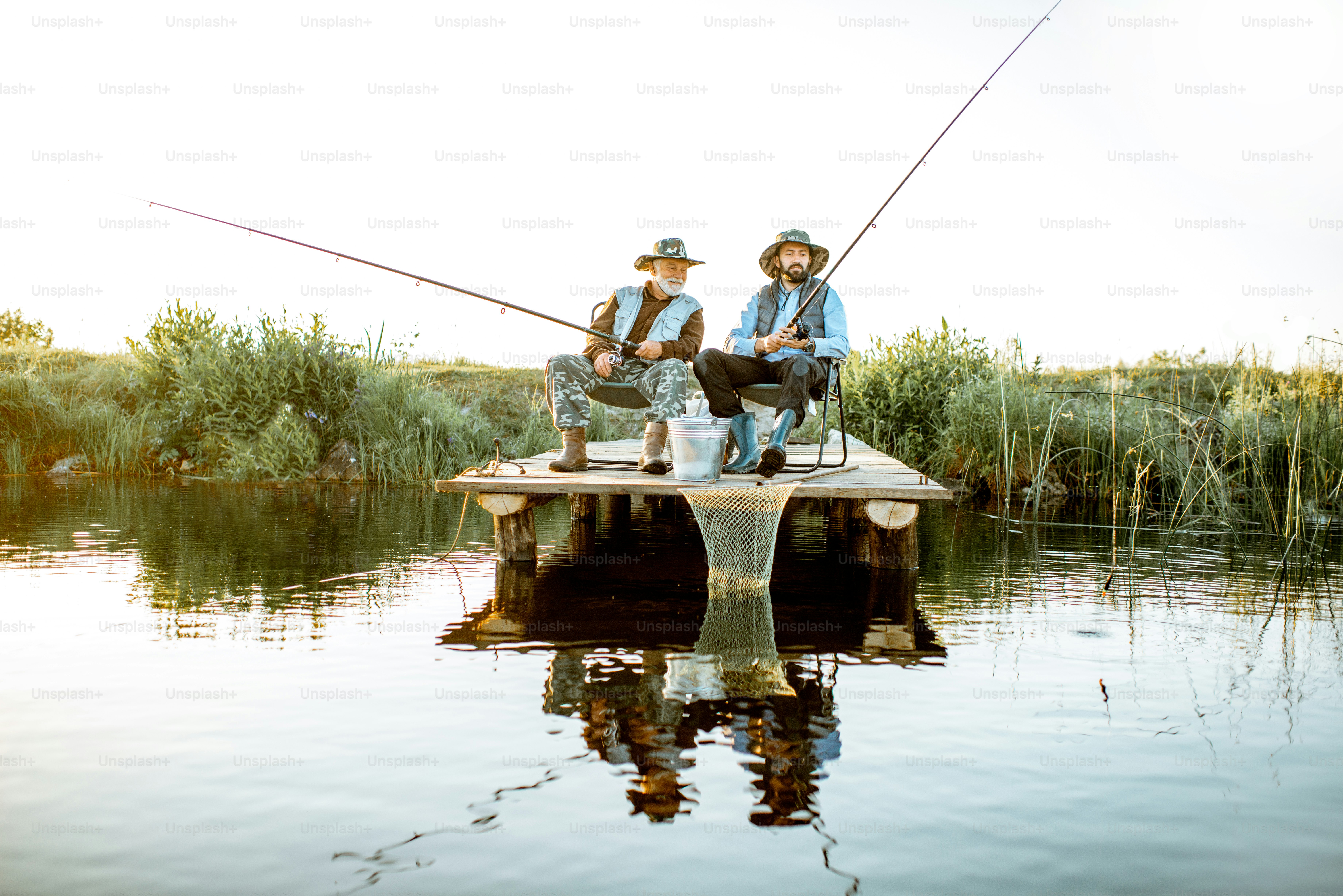 Avô com filho adulto pescando juntos no cais de madeira durante a luz da manhã. Vista do lado do lago