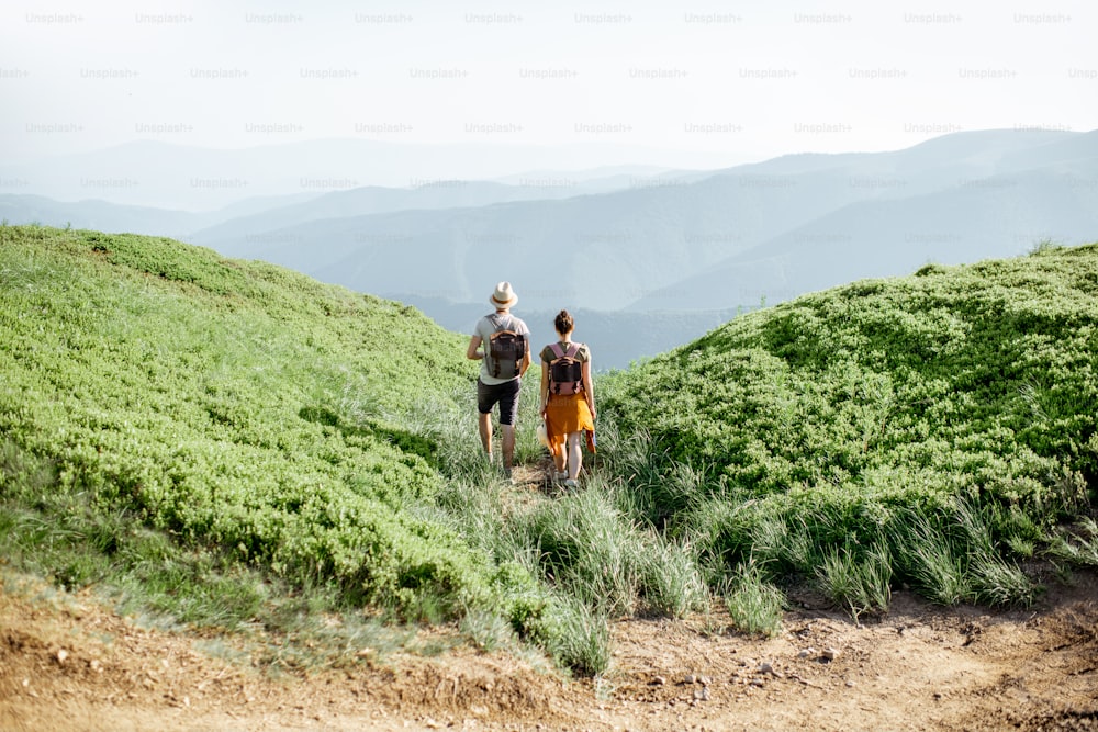 Beautiful couple walking with backpacks on the green meadow, while traveling high in the mountains during the summer time. Rear view