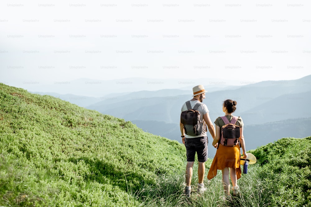 Schönes Paar, das mit Rucksäcken auf der grünen Wiese spazieren geht, während es im Sommer hoch in den Bergen unterwegs ist. Rückansicht