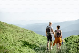 Beautiful couple walking with backpacks on the green meadow, while traveling high in the mountains during the summer time. Rear view