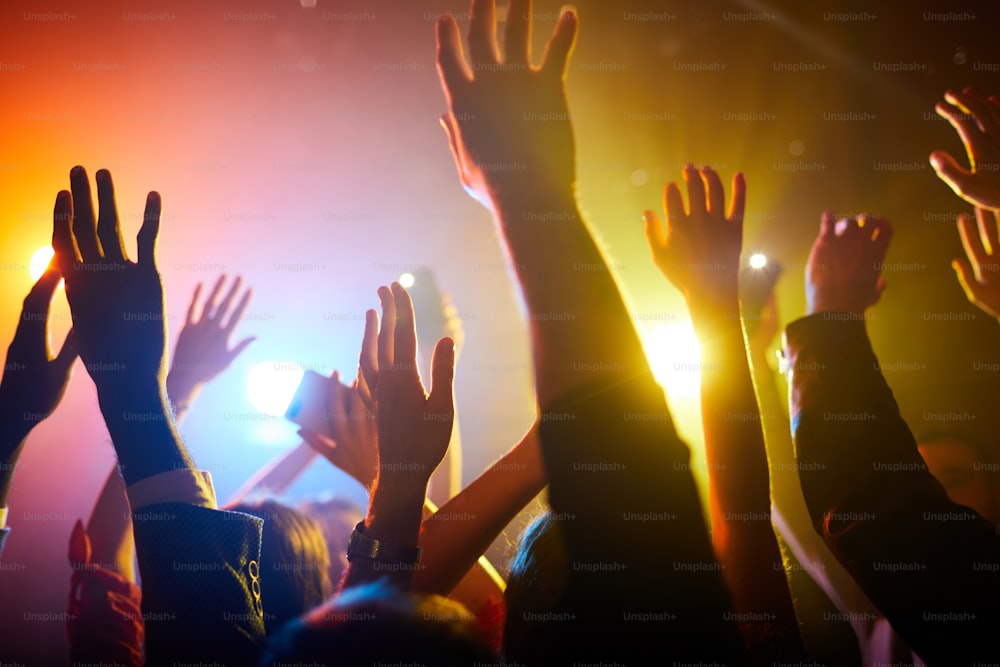 Group of unrecognizable people standing in colorful light and waving hands in air during performance of musician