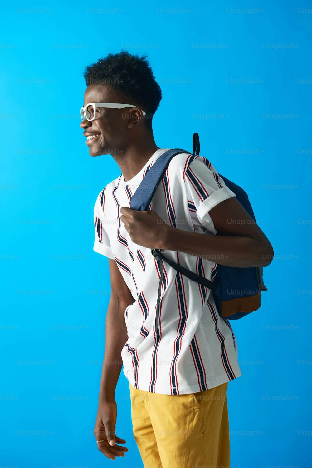 Profile view of young Aframerican student wearing white glasses and striped t-shirt. He smiling while standing on blue background