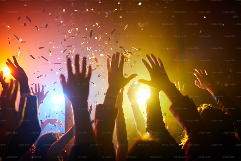 Grupo de pessoas irreconhecíveis em pé sob confetes e levantando as mãos em apresentação musical