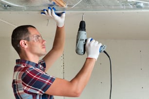 光沢のあるアルミホイルで断熱された天井の電気ドライバーを使用して、乾式壁の吊り天井を金属フレームに固定するゴーグルの若い男性。リノベーション、建設、自分でコンセプトをやる。