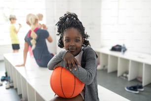 Pequeño jugador de baloncesto. Colegiala feliz sosteniendo su pelota de baloncesto sentada en el banco esperando su lección de deportes.