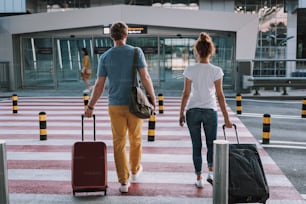 Rückansicht in voller Länge Porträt von Herrn und Dame, die ihre Trolley-Taschen tragen und Fußgängerüberwege benutzen. Sie gehen zum Eingang des Flughafenterminals