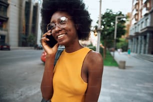 Waist up portrait of smiling afro-american female in glasses standing on the street and using mobile phone. Copy space in left side