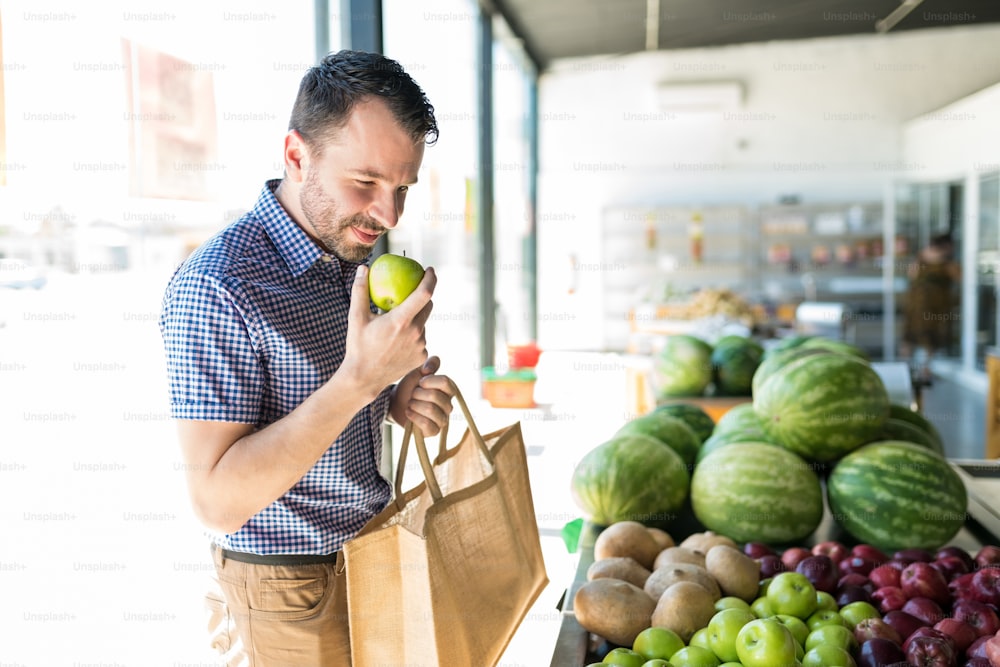 Hombre oliendo manzana verde orgánica mientras está parado en la tienda de comestibles