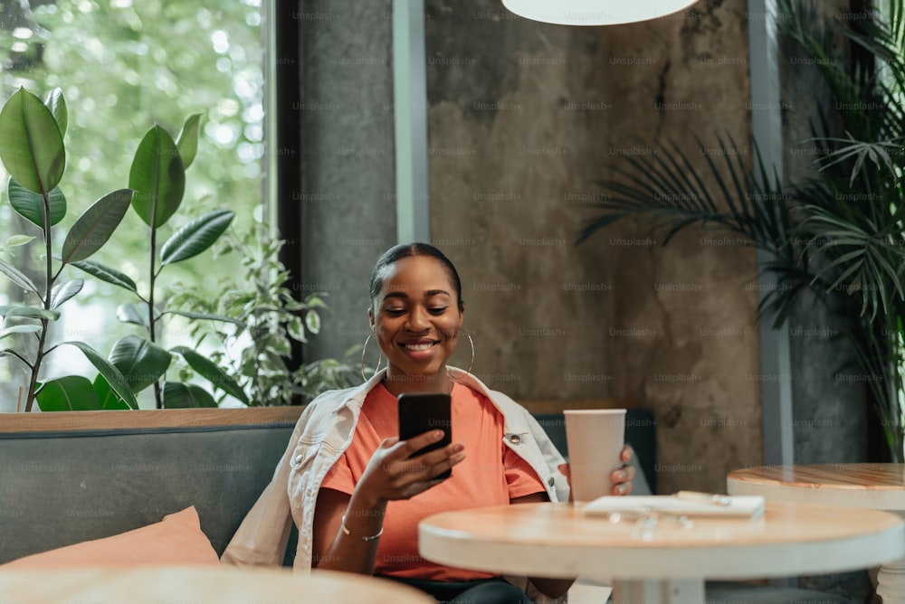 Cheerful young afro-american woman sitting inside cafe behind wooden table with mobile phone