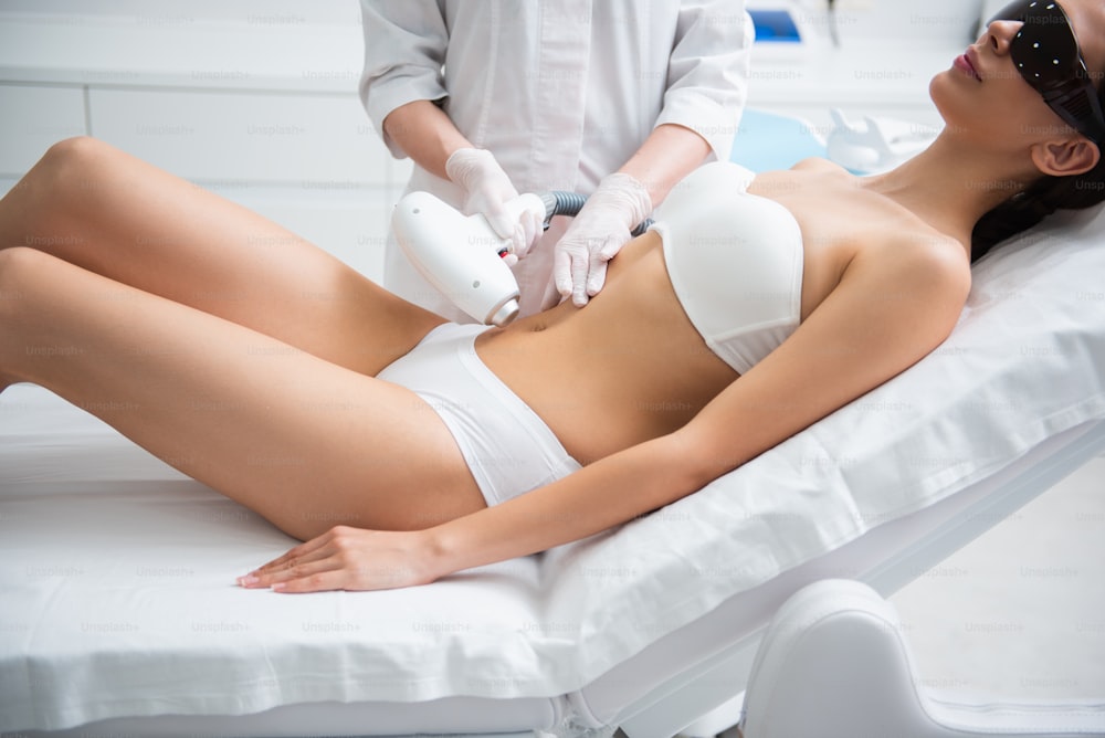 Aesthetic body treatment. Side on portrait of young slender relaxed woman in white underwear having hair removal procedure of belly zone
