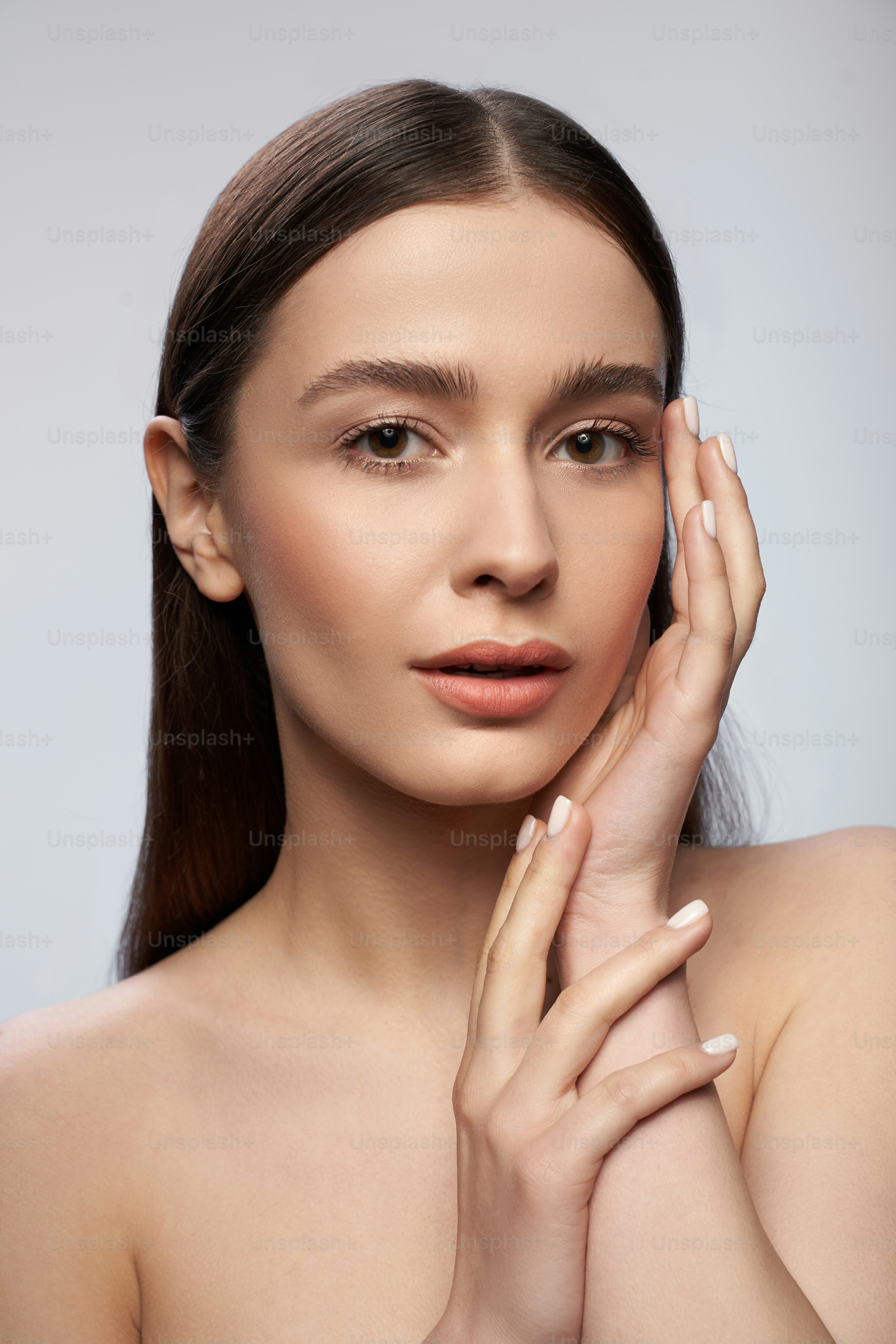 Calm young lady standing isolated against the light gray background while touching the cheek with her hand