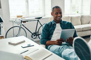 Handsome young African man in shirt using digital tablet and smiling while sitting in the office