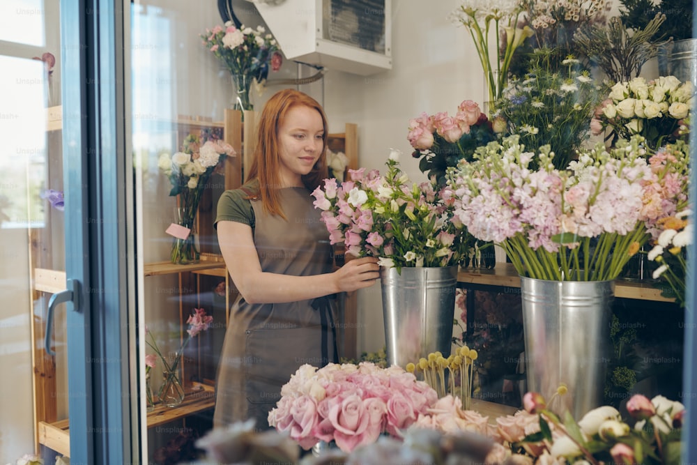 Content beautiful young redhead flower shop worker in apron standing in flower refrigerator and checking flowers in metal bucket