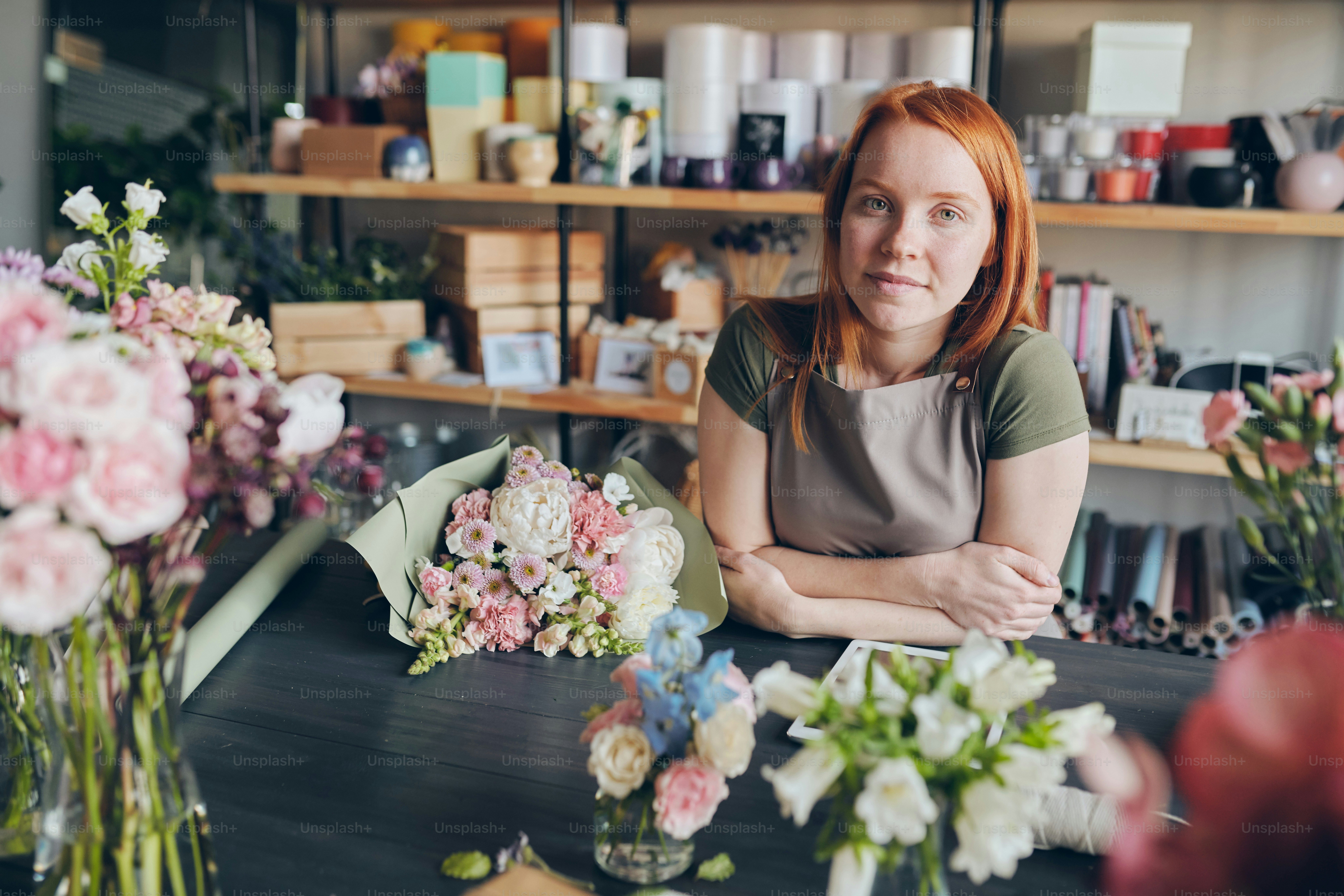 現代の花屋で様々な花を持つカウンターに立つエプロンを着た赤毛の花屋のポートレート