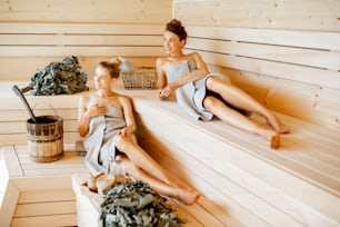 Two young girlfriends relaxing in the sauna, lying on the wooden benches with bucket and bath brooms