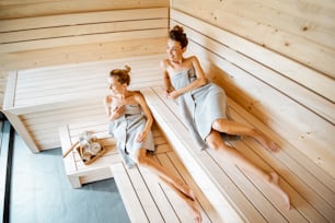 Dos jóvenes amigas relajándose en la sauna, acostadas en los bancos de madera con cubo y escobas de baño