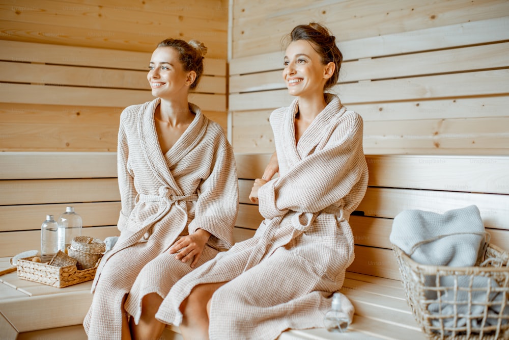 Zwei junge schöne Freundinnen in Bademänteln sitzen zusammen, während sie sich in der Luxussauna entspannen