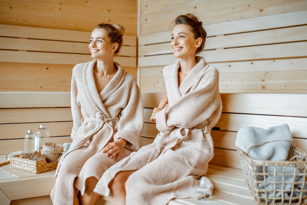 Two young beautiful girlfriends in bathrobes sitting together while relaxing in the luxury sauna